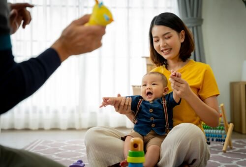A woman is holding a baby and smiling. The baby is playing with a toy. The scene is warm and happy
